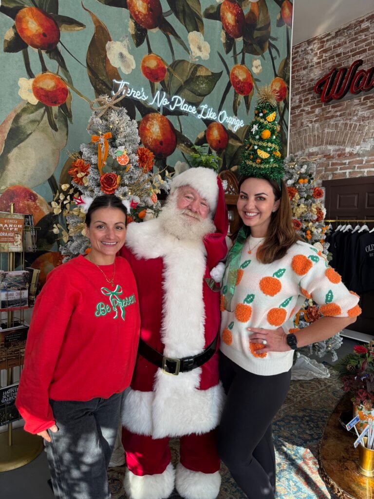 Santa Claus poses with two women in front of a festive orange-themed holiday backdrop featuring decorated Christmas trees and a neon sign that reads “There’s No Place Like Orange.