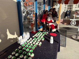 Santa Claus poses with a family beside a table filled with green-frosted cupcakes and white cups, outside a festive storefront during a holiday event.