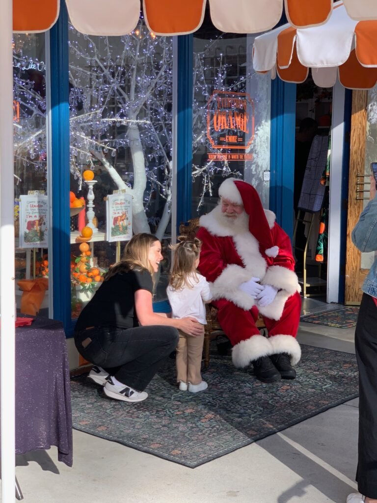 Santa Claus interacts with a young child and their guardian in front of a winter-themed storefront decorated with white lights.