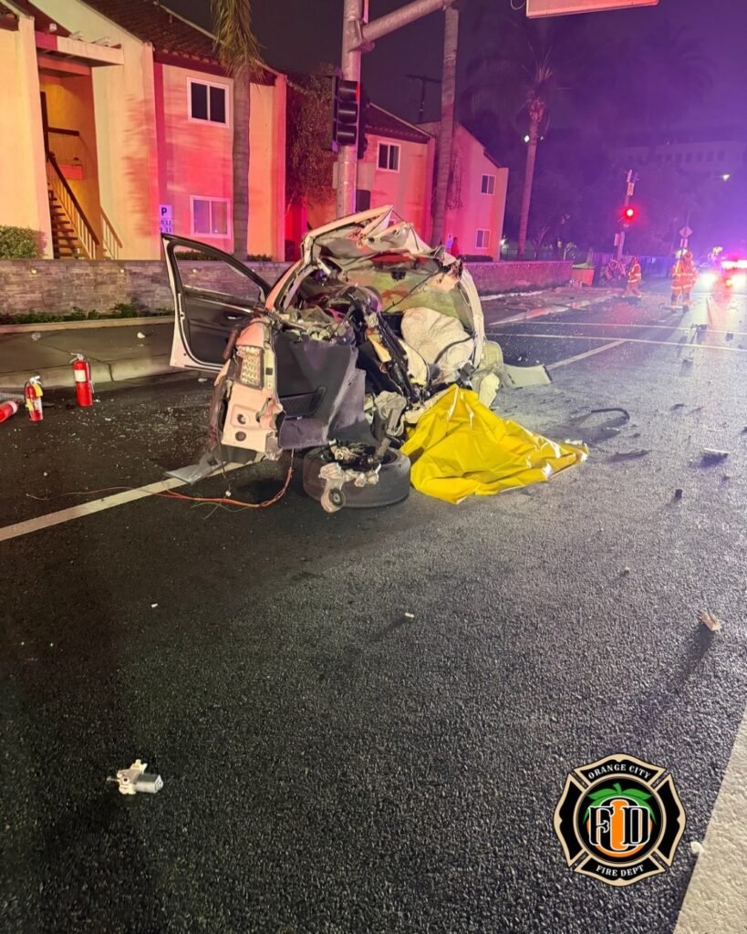 Severely damaged vehicle with yellow tarp and emergency crews at fatal crash scene in Orange.