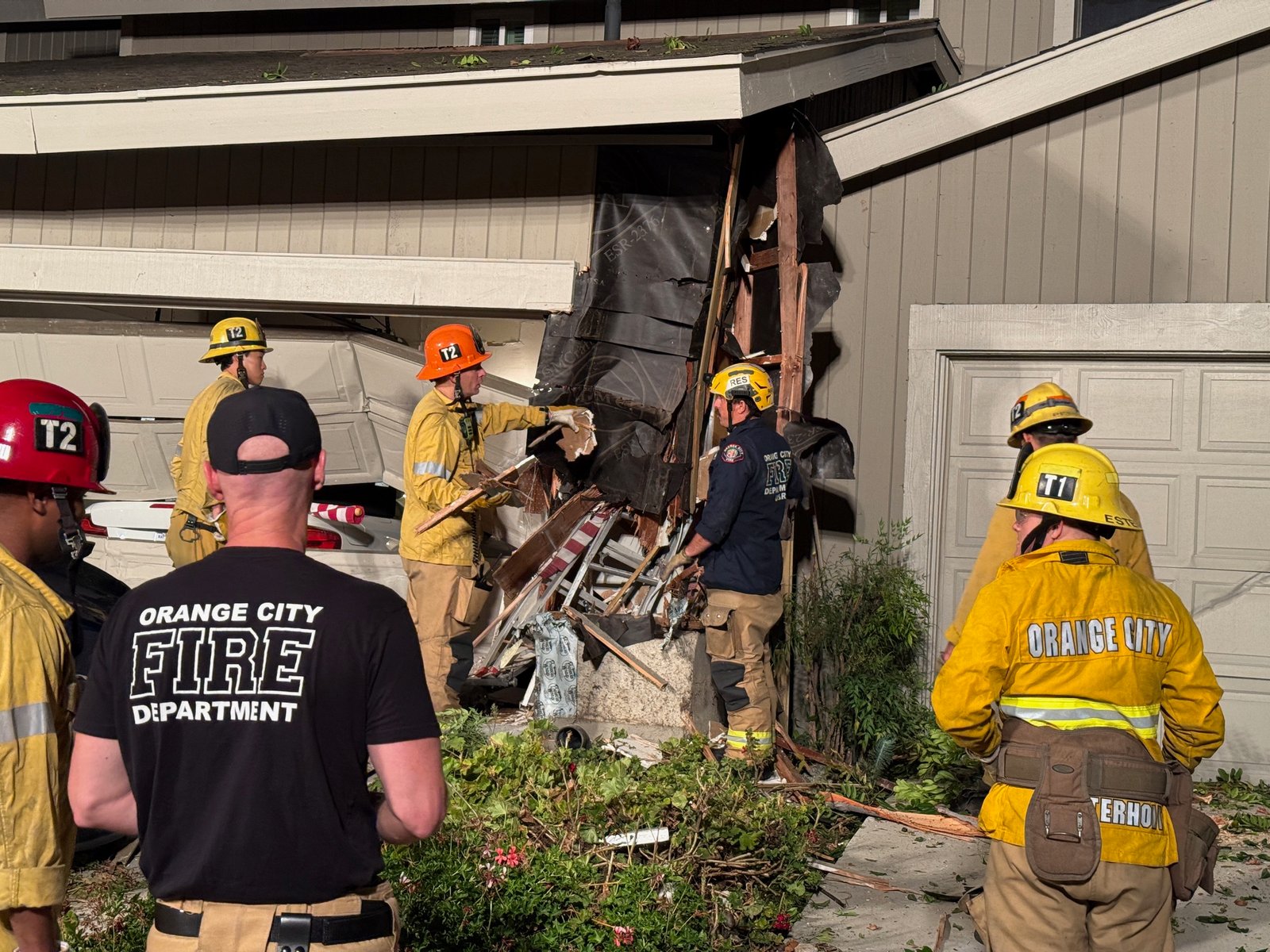 Orange firefighters inspect a torn-open exterior wall and debris beside a home's garage after the crash.