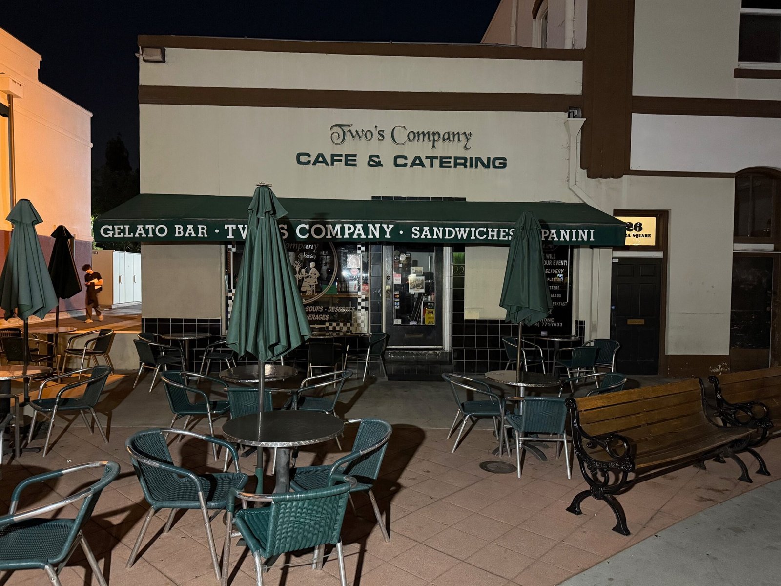 Outdoor tables, chairs and closed umbrellas outside Two's Company Cafe & Catering in the Orange Plaza at night.