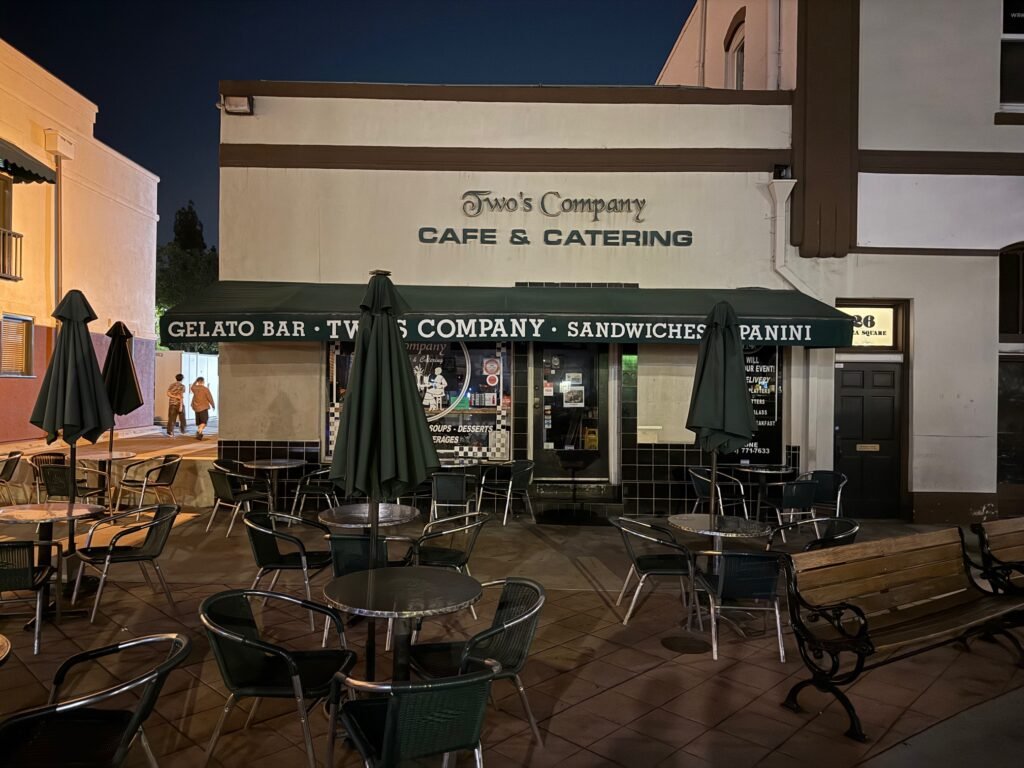 Sidewalk dining tables and a wooden bench in front of Two's Company Cafe & Catering.