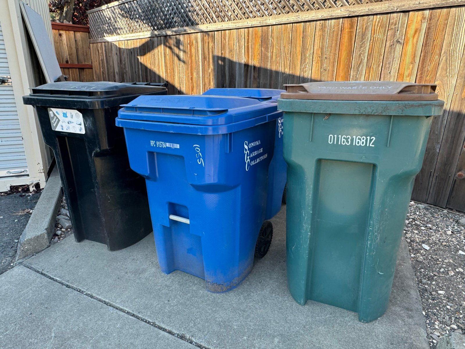 Black, blue and green waste carts beside a wooden fence in Sonoma, California.